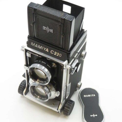A vintage Mamiya C220 twin-lens reflex camera with a black and silver design, featuring two prominent lenses and a waist-level viewfinder up. Beside the camera is a matching lens cap with the Mamiya logo.