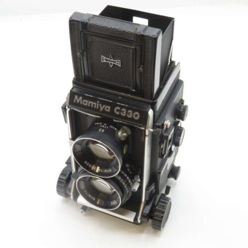 A vintage Mamiya C330 twin-lens reflex camera with a black body and dual lenses, featuring various knobs and dials for manual adjustments, set on a white background.