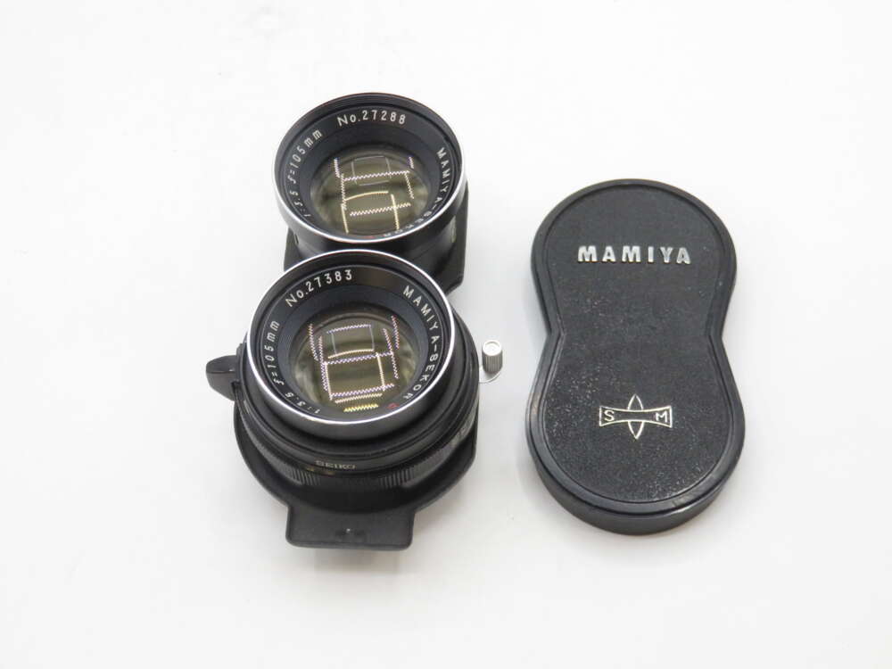 IMG_1778 A pair of Mamiya camera lenses with a matching lens cap on a white background. The lenses are labeled as 105mm f/3.5, and the lens cap is black, marked with the Mamiya logo.