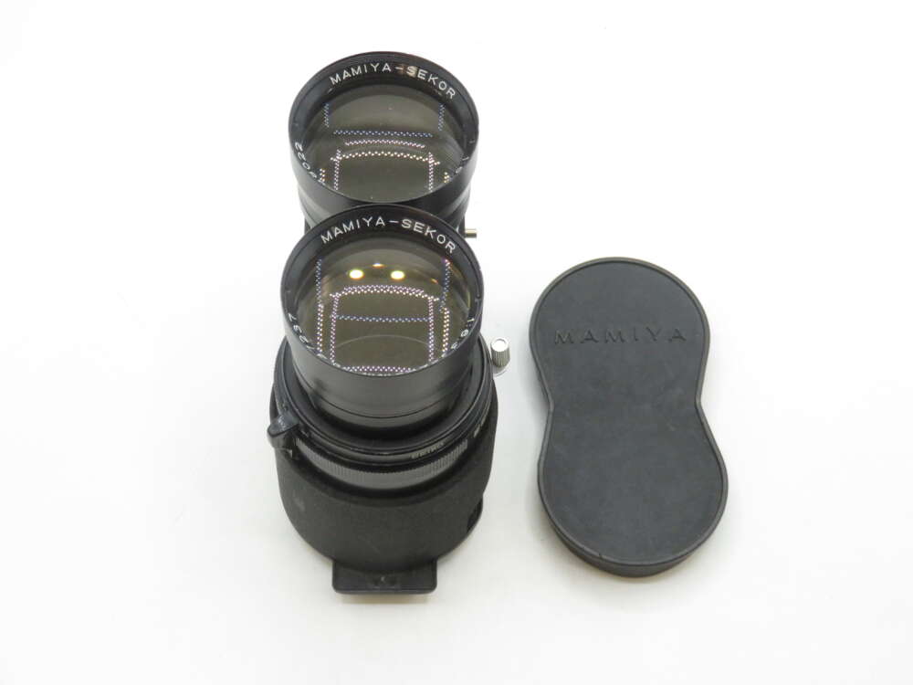 IMG_1779 A pair of Mamiya-Sekor camera lenses stacked vertically, with a black lens cap bearing the Mamiya logo placed beside them on a white surface.