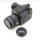 A Pentax camera with a detachable lens is placed on a white surface, with its lens cap lying next to it.