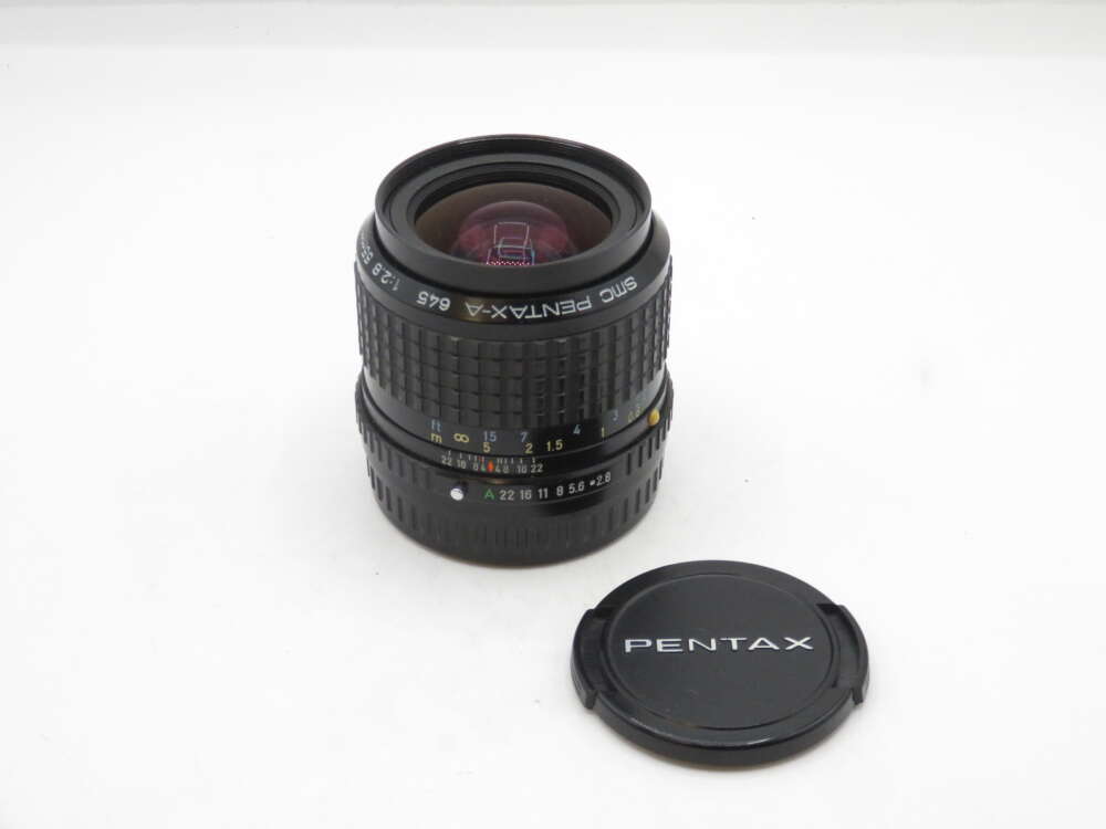 IMG_1796 A Pentax-A 645 1:2.8 45mm camera lens with a detachable black lens cap beside it, placed on a plain white surface.