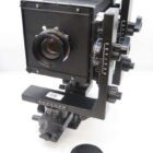 A large format camera with bellows, various adjustment knobs, and a lens at the front. The camera is mounted on a tripod head with precise calibration markings. A lens cap is placed nearby on a white surface.