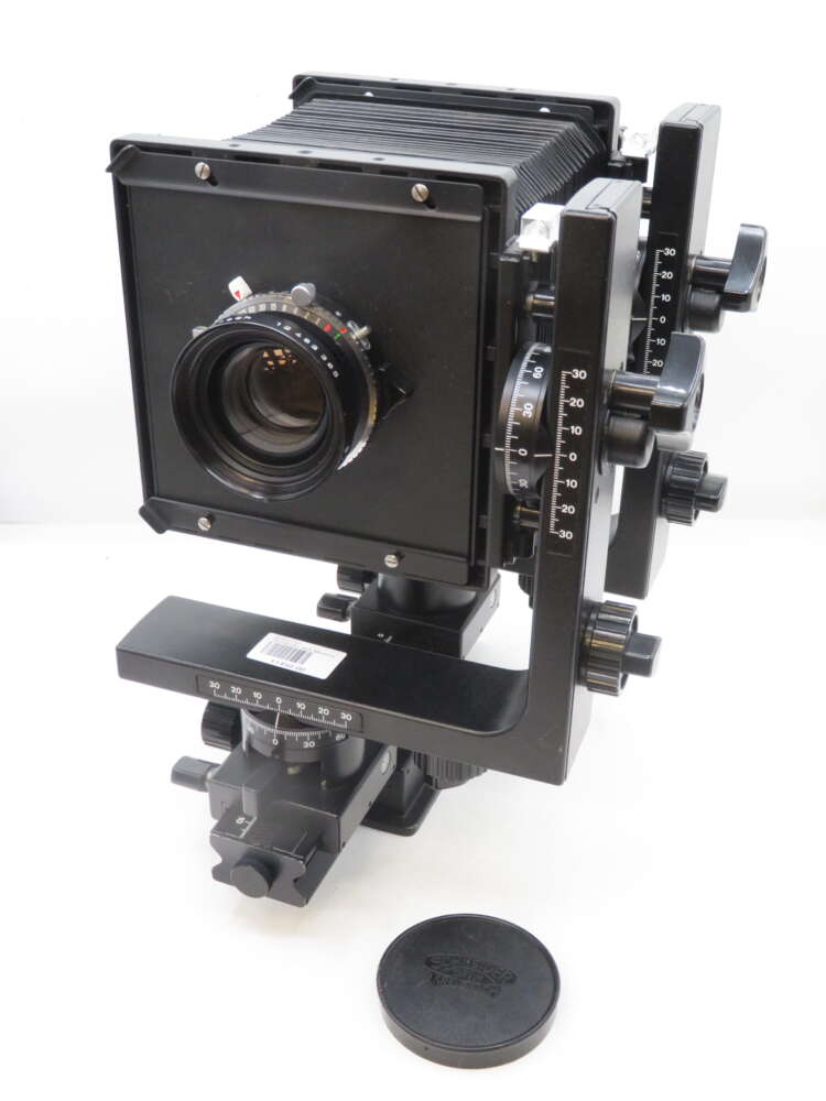 IMG_1815 A large format camera with bellows, various adjustment knobs, and a lens at the front. The camera is mounted on a tripod head with precise calibration markings. A lens cap is placed nearby on a white surface.