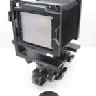 A large format camera with a bellows design, featuring adjustment knobs and a ground glass focusing screen, set on a white background. A lens cap rests nearby.