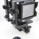 A black large format camera with a bellows design, mounted on a tripod head with various adjustment knobs and scales.