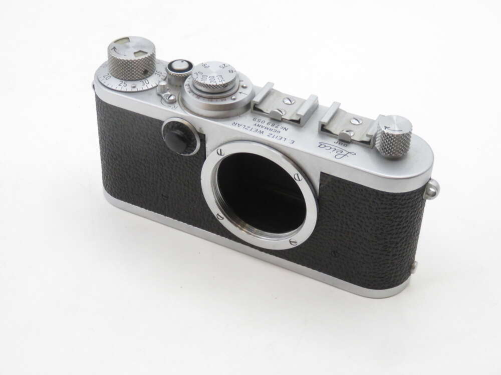 IMG_1824 A vintage rangefinder camera body with a black textured exterior and silver metal trim, featuring dials and knobs on the top, with an open lens mount at the front.