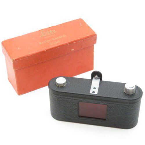 A vintage Leitz Kepler-Aparat E 3/4 optical device in black is displayed next to its original orange box. The device has a rectangular shape with two metallic knobs on top and a viewing lens, embodying a classic design.