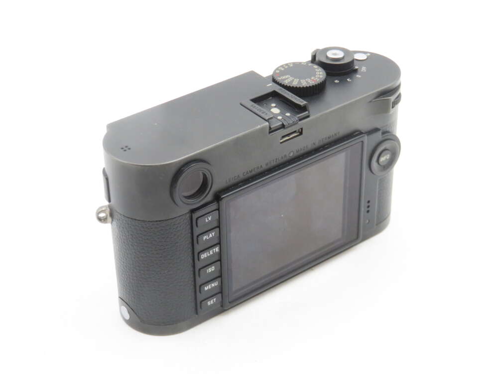 A black Leica digital camera shown from the back, featuring a large LCD screen and several control buttons on the left side of the screen. The top of the camera has a visible dial and a hot shoe mount. The body has a textured grip on one side.