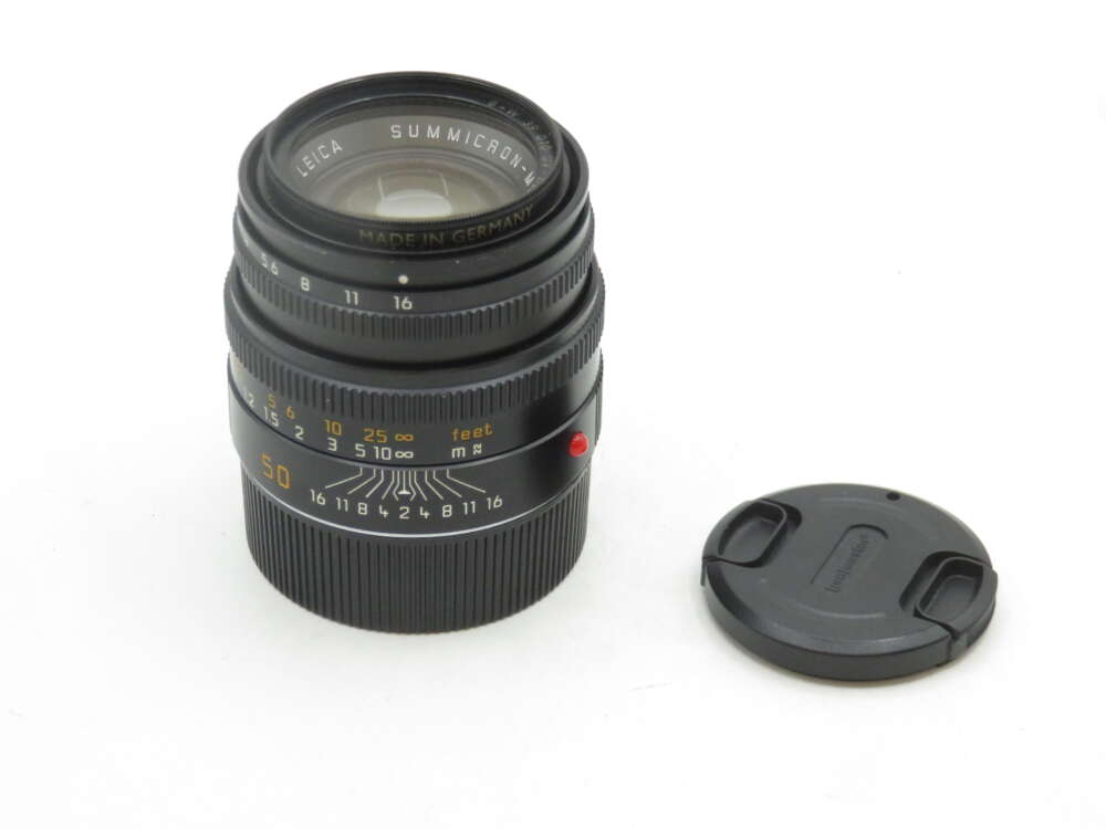 A black Leica Summicron 50mm camera lens, displayed upright on a white surface, with its lens cap placed next to it. The lens has markings for aperture and focus distances.