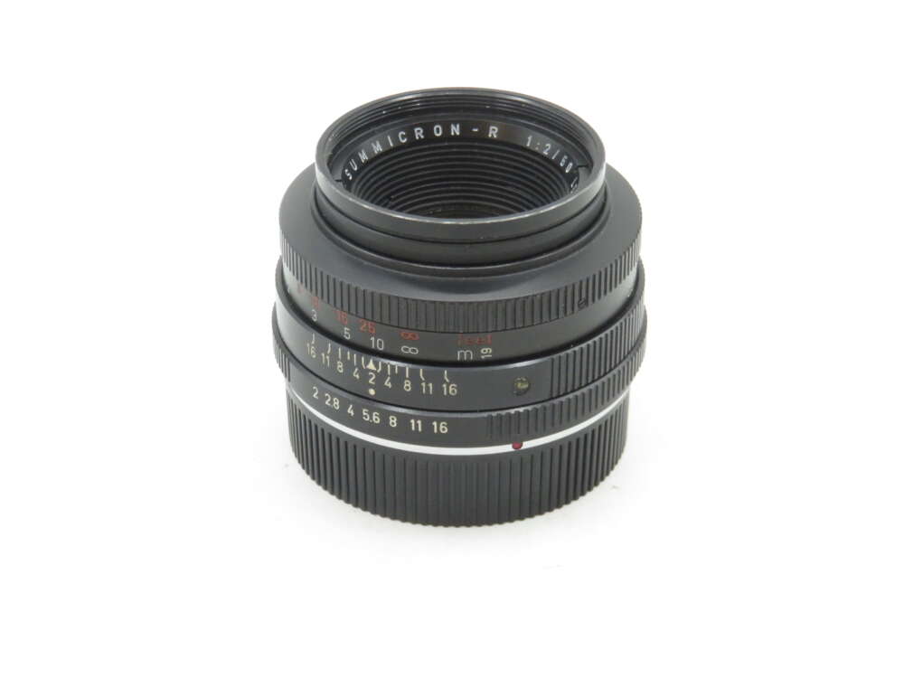 A black camera lens with multiple engraved markings, including "SUMMICRON-R 1:2/50," featuring an aperture and focus ring, set against a plain white background.