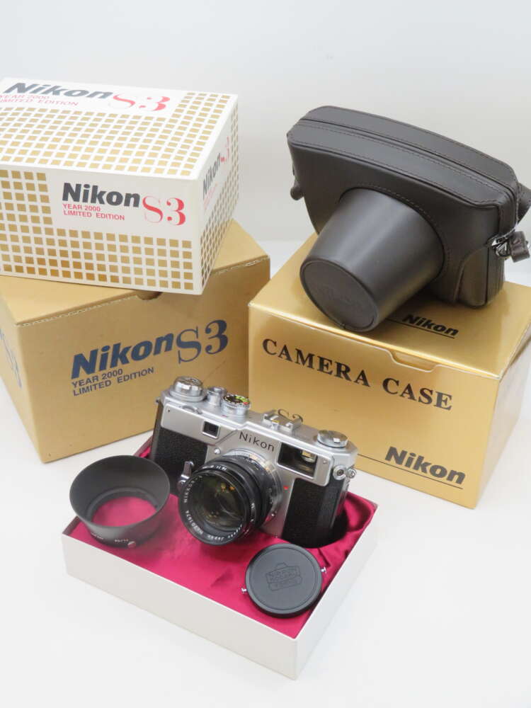 IMG_2016 A Nikon S3 Year 2000 Limited Edition camera is displayed alongside its original boxes and a camera case. The camera, featuring a classic silver and black design, is set on a red velvet-lined box with a lens hood and lens cap nearby. Prominent boxes indicate the product as a limited edition item.