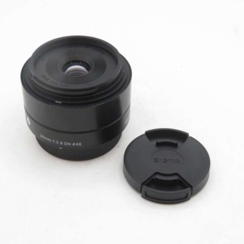 A Sigma 30mm f/2.8 DN lens placed on a white surface next to its lens cap.