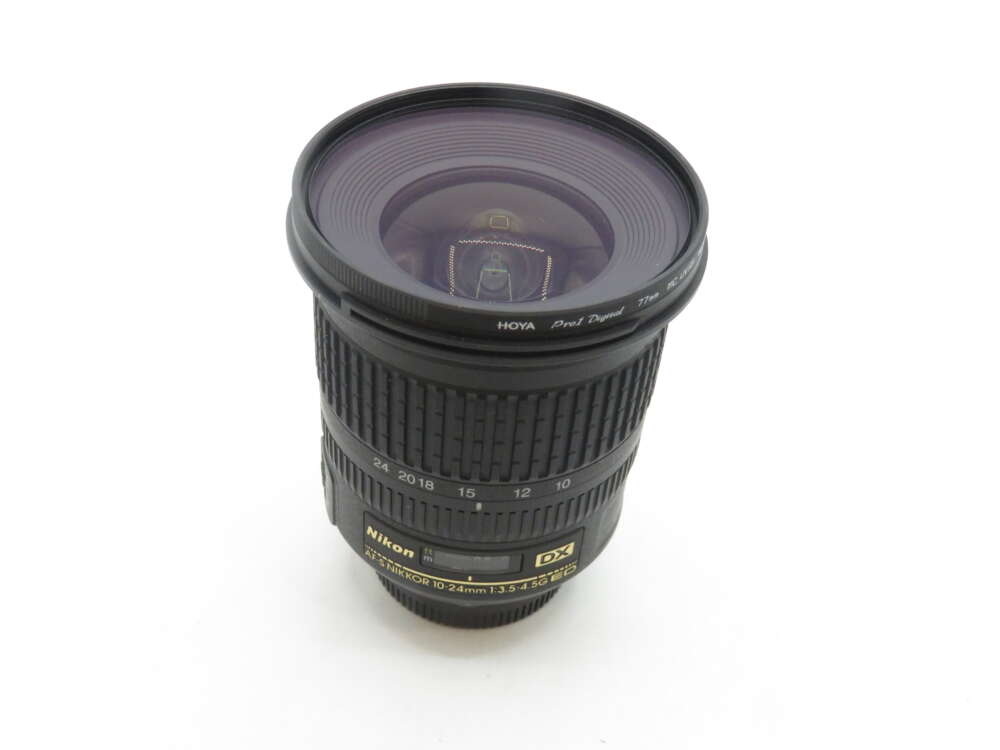 IMG_2160 Nikon DX AF-S NIKKOR 10-24mm f/3.5-4.5G ED black camera lens with visible focus and zoom rings, and a Hoya Pro1 Digital 77mm lens filter attached, standing upright against a white background.