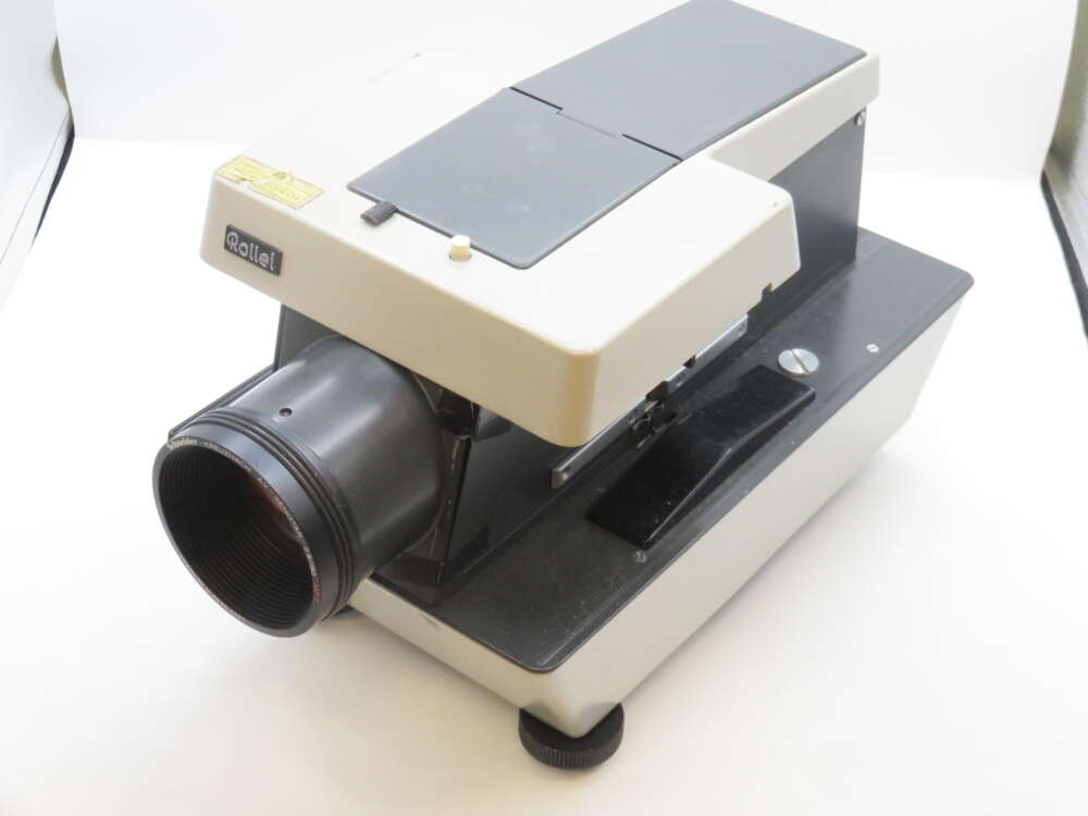 IMG_2207 A vintage Rollei slide projector with a black and beige body, featuring a prominent lens at the front and a large, flat top surface. The projector is positioned on a white background, highlighting its retro design and mechanical components.