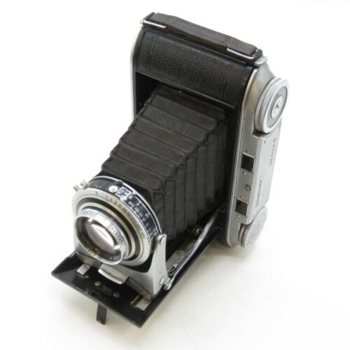 A vintage folding camera with a bellows design, featuring a metallic lens and knobs on the side, set against a plain white background.