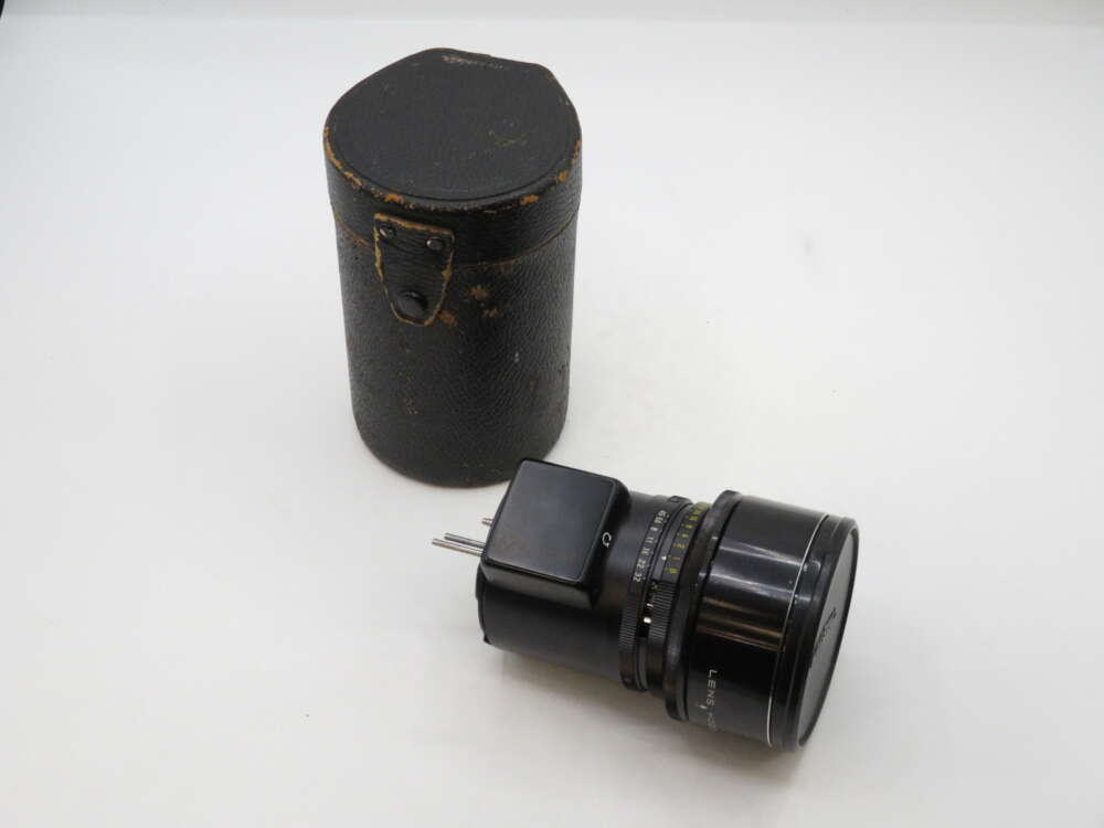 A vintage camera lens with a cap is lying next to a cylindrical black leather case with a lid, placed on a white surface.