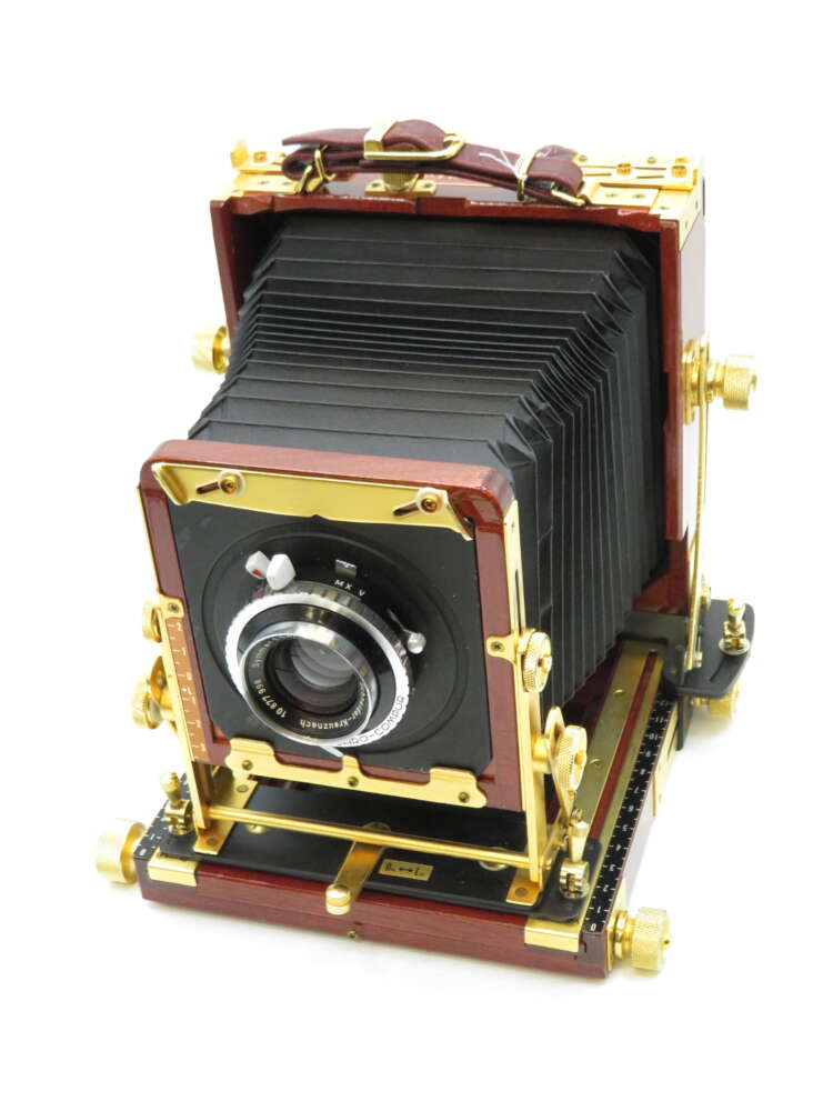 A vintage large format camera with a dark bellows design, polished wood frame, and gold-tone metal fittings, featuring a prominent lens at the front.