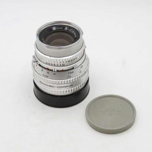 A vintage camera lens with a silver exterior and various control rings is next to a grey lens cap.
