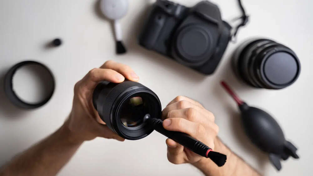 Hands cleaning a camera lens with a brush, surrounded by camera equipment on a white surface.
