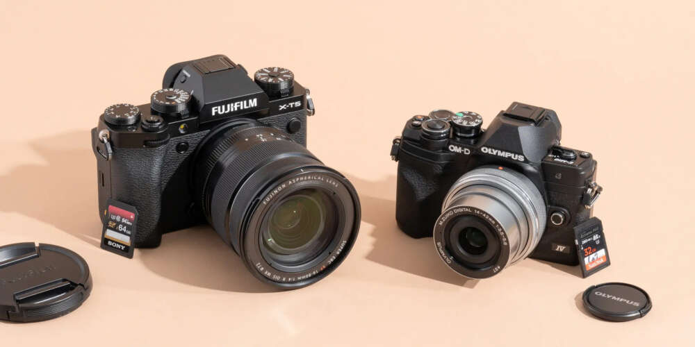 Two black digital cameras, a Fujifilm X-T5 with a large lens and memory card inserted, and an Olympus camera, both on a beige surface.
