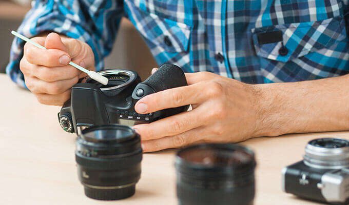 A person cleaning the sensor of a DSLR camera with a swab, surrounded by multiple camera lenses on a table.