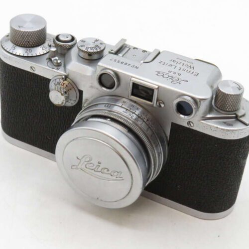 A vintage Leica camera with a black textured body, silver dials, and a lens cap displaying the Leica logo.