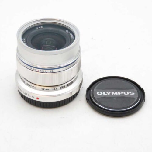 A silver Olympus camera lens with a black lens cap featuring the Olympus logo next to it.