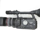 A Canon professional video camera with a large lens and handle, viewed from the side against a white background.