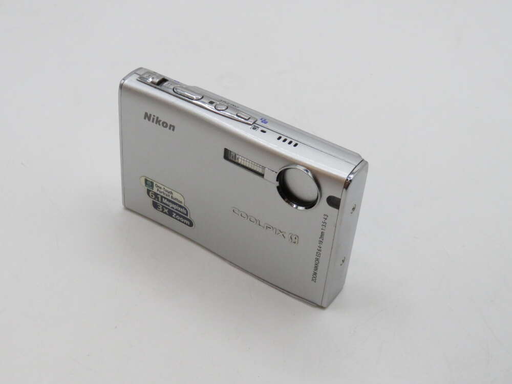 IMG_3433 Silver Nikon Coolpix digital camera on a white surface, featuring a lens, flash, and various buttons on top.