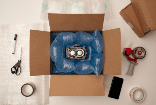 A camera is nestled in blue air cushions inside an open cardboard box, surrounded by packing materials, a phone, and scissors.