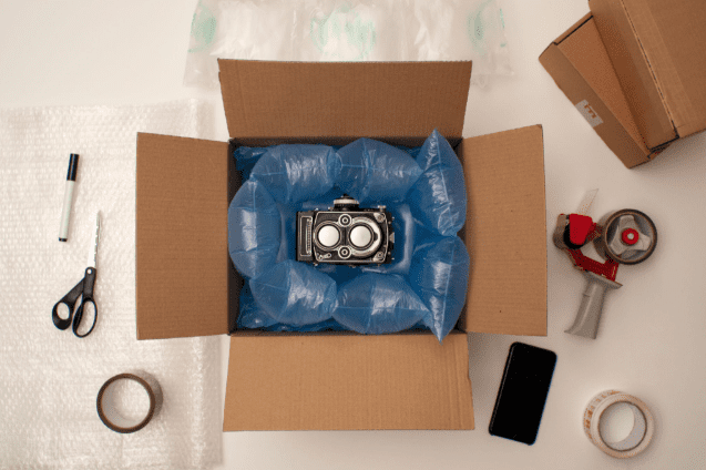 A camera is nestled in blue air cushions inside an open cardboard box, surrounded by packing materials, a phone, and scissors.