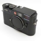 Black Leica M9 camera with a textured grip, featuring a red Leica logo and a lens cap on a neutral background.
