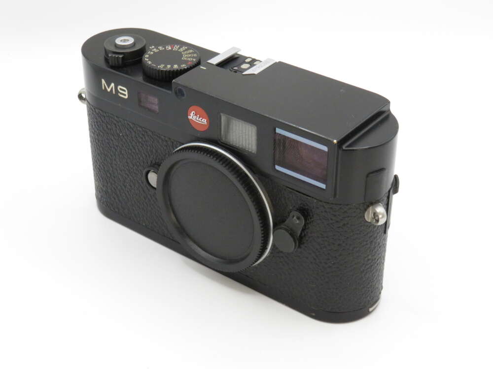 IMG_3705 Black Leica M9 camera with a textured grip, featuring a red Leica logo and a lens cap on a neutral background.