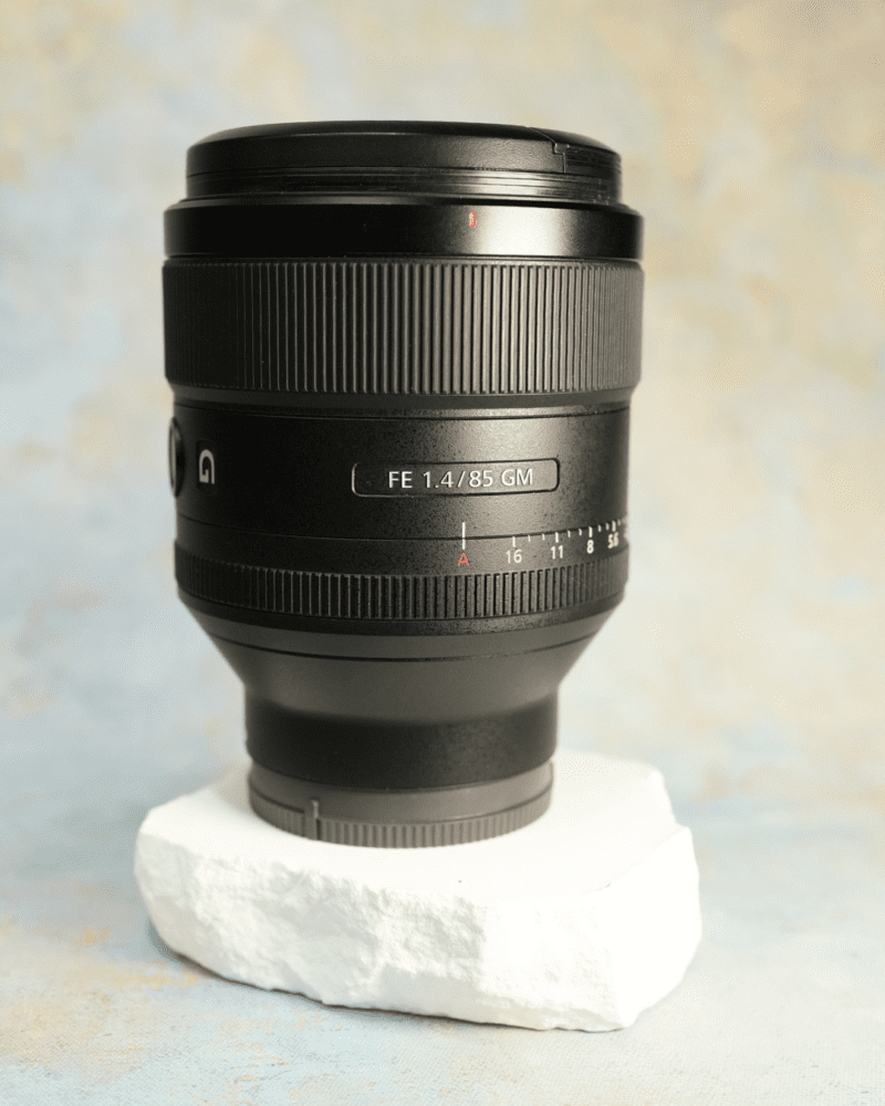 A black camera lens marked as FE 1.4/85 GM, resting on a piece of white styrofoam against a soft, multicoloured background.