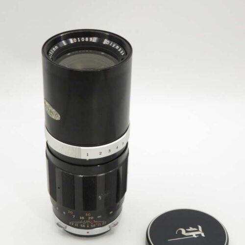 A black vintage camera lens with a silver ring, featuring visible numbers and a detached lens cap with a logo next to it.
