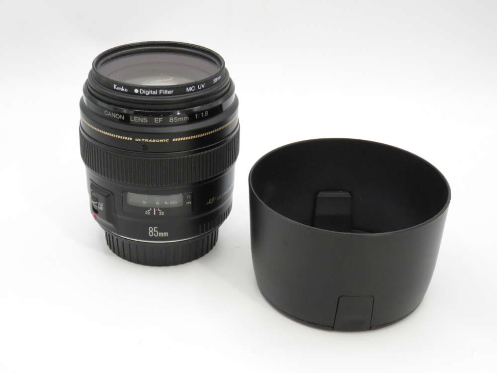 IMG_3917 Canon EF 85mm f/1.8 lens with a lens hood placed beside it on a plain background.