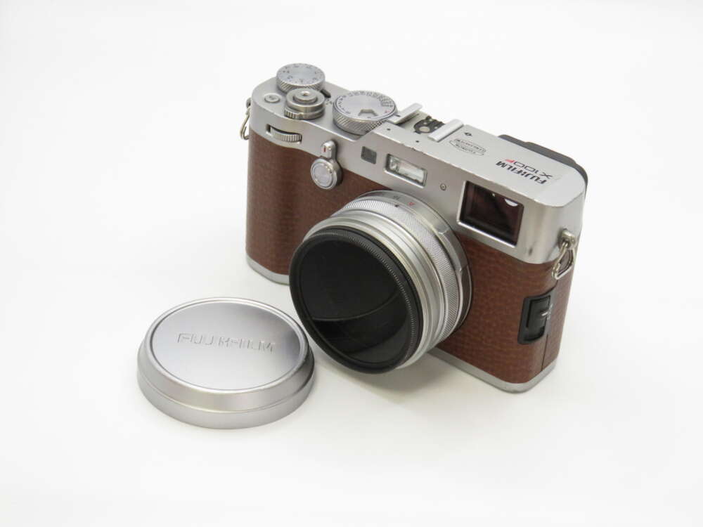 IMG_3994 A brown and silver Fujifilm camera with a retro design next to its lens cap on a white surface.