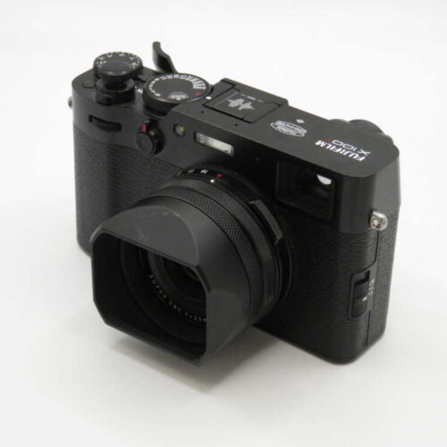 A black Fujifilm X100 camera with a textured grip, lens hood, and various control dials on a white surface.
