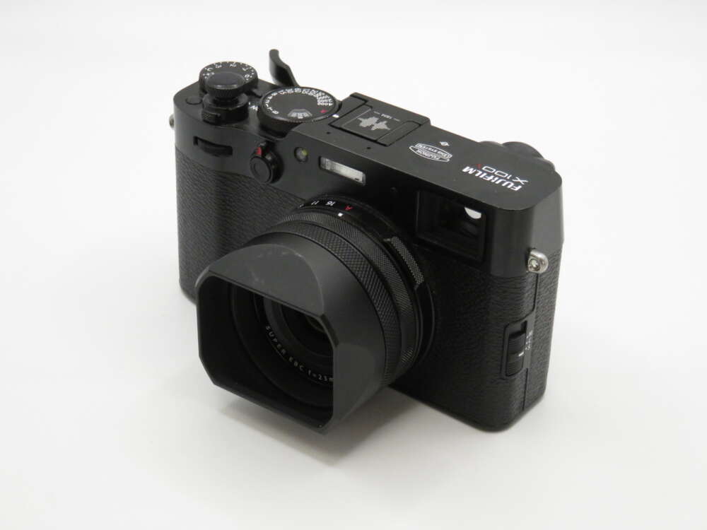 IMG_4132 A black Fujifilm X100 camera with a textured grip, lens hood, and various control dials on a white surface.