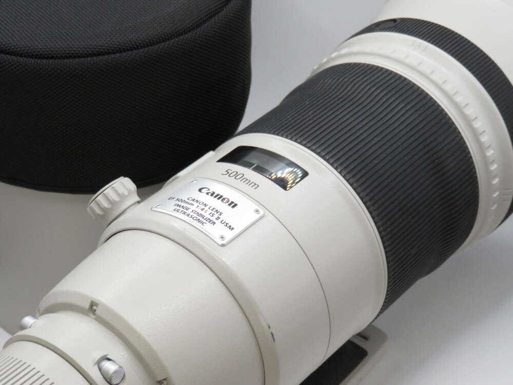 A white Canon 500mm telephoto lens with black detailing, set beside a matching cylindrical lens case.
