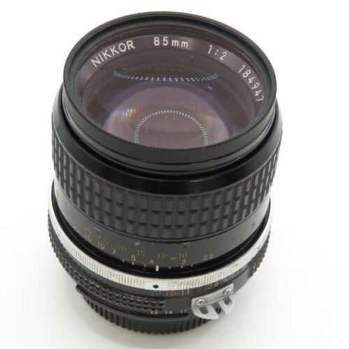 A camera lens with "Nikkor 85mm 1:2" markings on the front, featuring a focus ring and aperture adjustment.